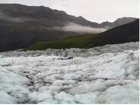 Personas en un paisaje con hielo y montañas, nubes bajas y vegetación en el fondo.
