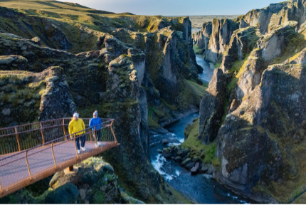 Dos personas caminando por un puente suspendido sobre un profundo cañón con ríos y paredes rocosas.