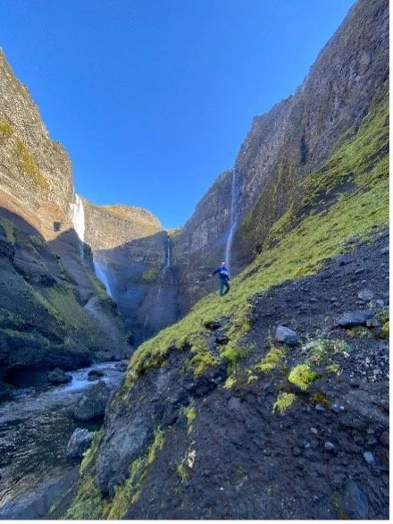 Persona explorando un cañón con cascadas, rodeada de paredes de roca cubiertas de musgo y un río en la base.