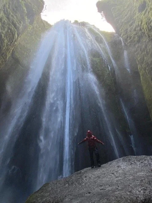 Persona con chaqueta roja frente a una cascada en un cañón con paredes verdes.