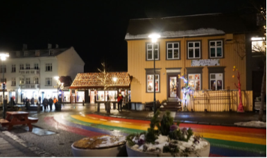 Escenario exterior con iluminación navideña, casas decoradas y una bandera de arcoíris en la calle, en un entorno urbano nocturno.