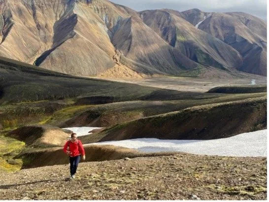 Persona caminando en un paisaje montañoso con laderas de colores y un río