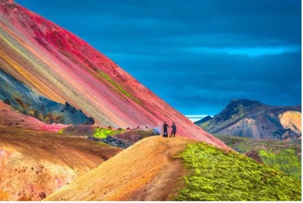 Dos personas observando un paisaje colorido con montañas de diferentes tonos y un cielo azul oscuro.