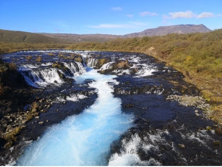 Río con cascadas en un paisaje natural con montañas y cielo azul.