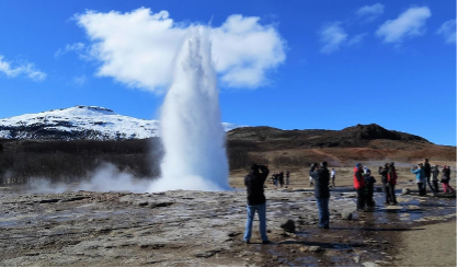 Vapor de geyser disparándose al aire con turistas observando cerca en un paisaje volcánico con montañas y cielo azul.