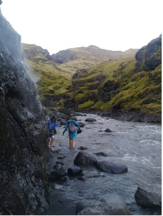 Dos personas con mochilas caminando por un río en un paisaje de montañas verdes.