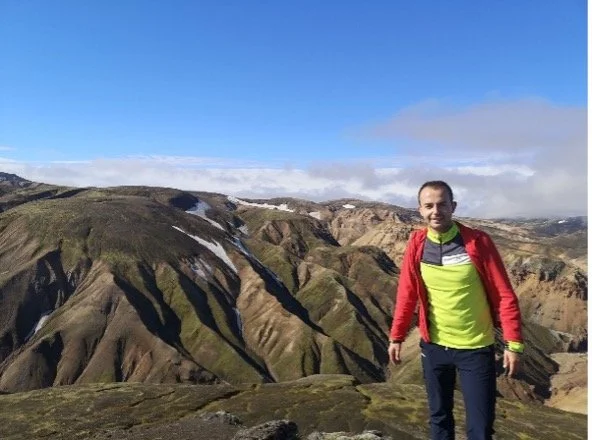 Hombre con ropa deportiva de pie en una montaña con formación rocosa y vegetación, bajo un cielo parcialmente nublado.