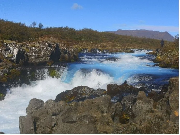 Un río con agua azul que atraviesa un paisaje de tierra y rocas, con colinas y montañas al fondo bajo un cielo despejado.