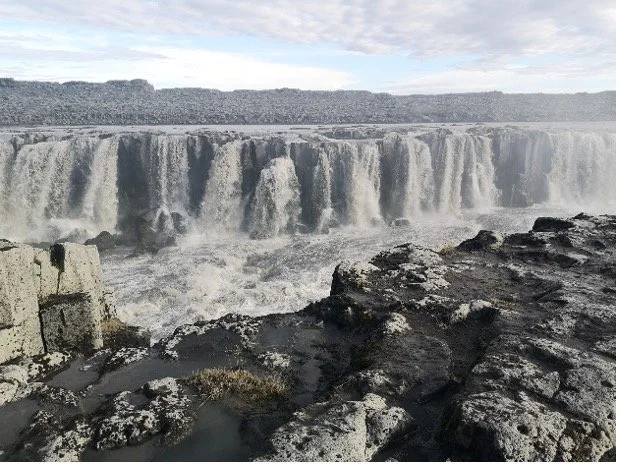Cascada de aguas caídas con rocas y forma de río en primer plano, cielo nublado al fondo.