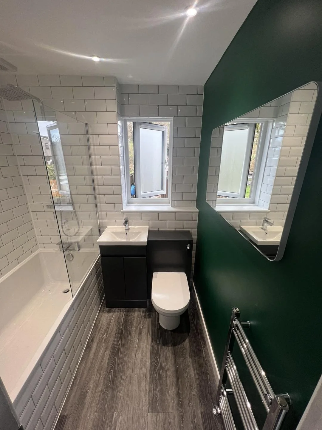 Small modern bathroom with white subway tile walls, a bathtub with a glass shower screen, small sink with black cabinet, toilet, two open windows, a large wall mirror, and a chrome towel radiator.