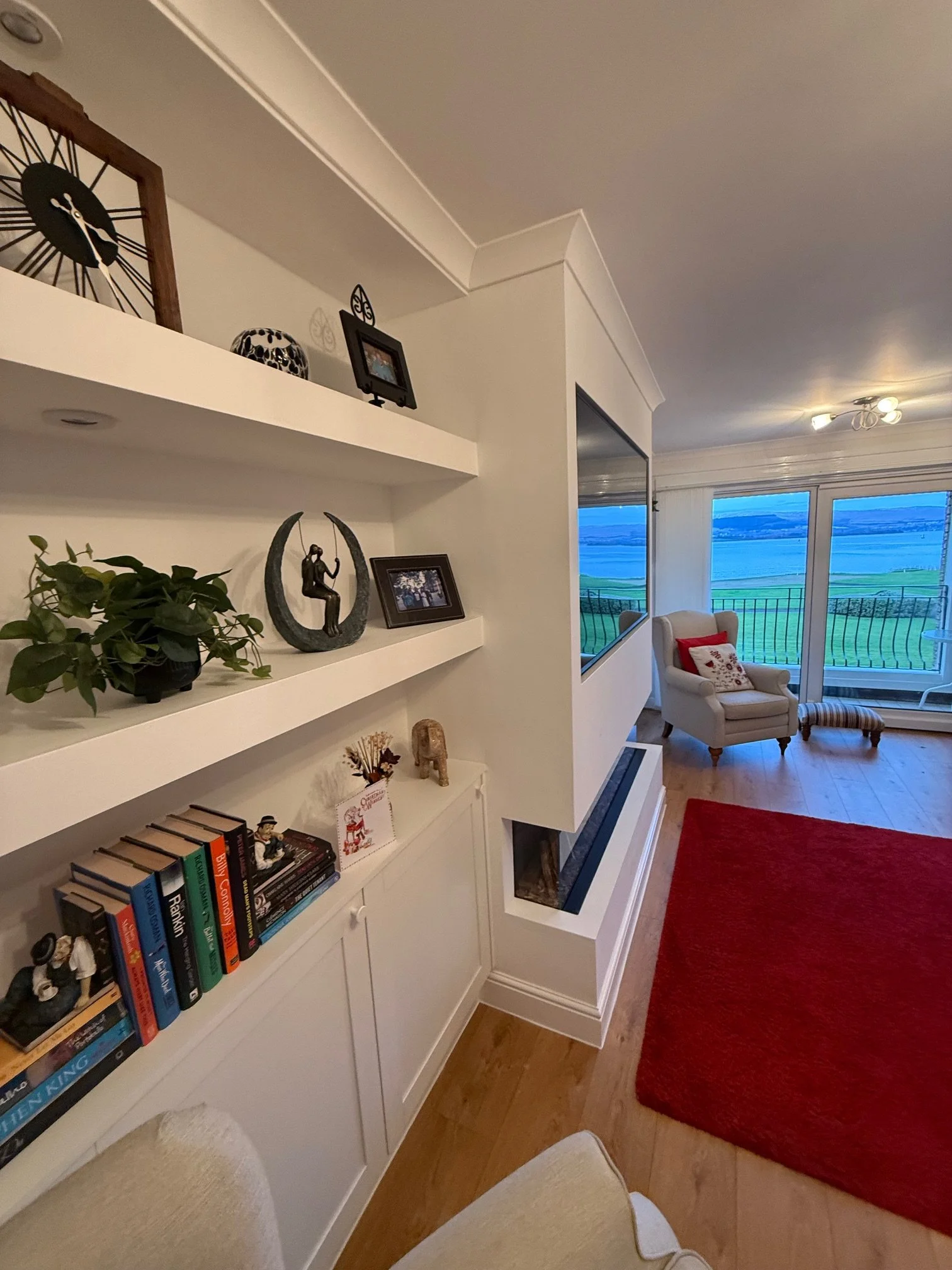 Living room with a view of a body of water outside large windows, featuring a white armchair with a red and white pillow, a small footstool, a red rug, and a built-in bookshelf with décor and books.
