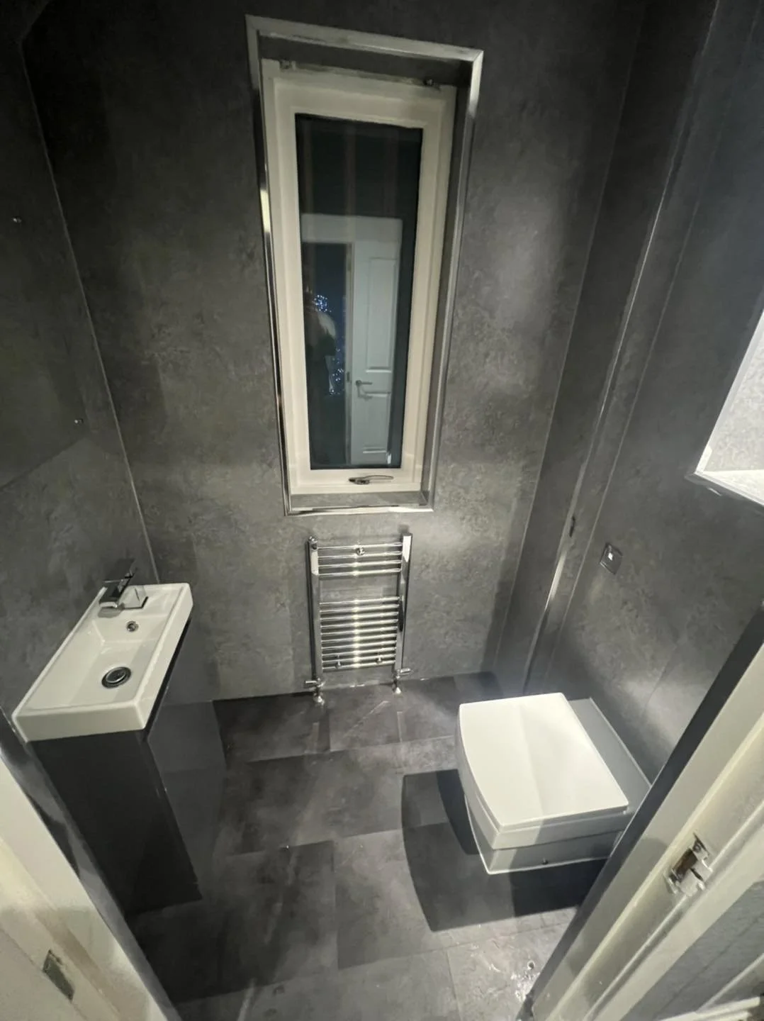 Small modern bathroom with gray tiled walls and floor, a compact white sink on a black pedestal, a toilet, a vertical window, and a heated towel rail.