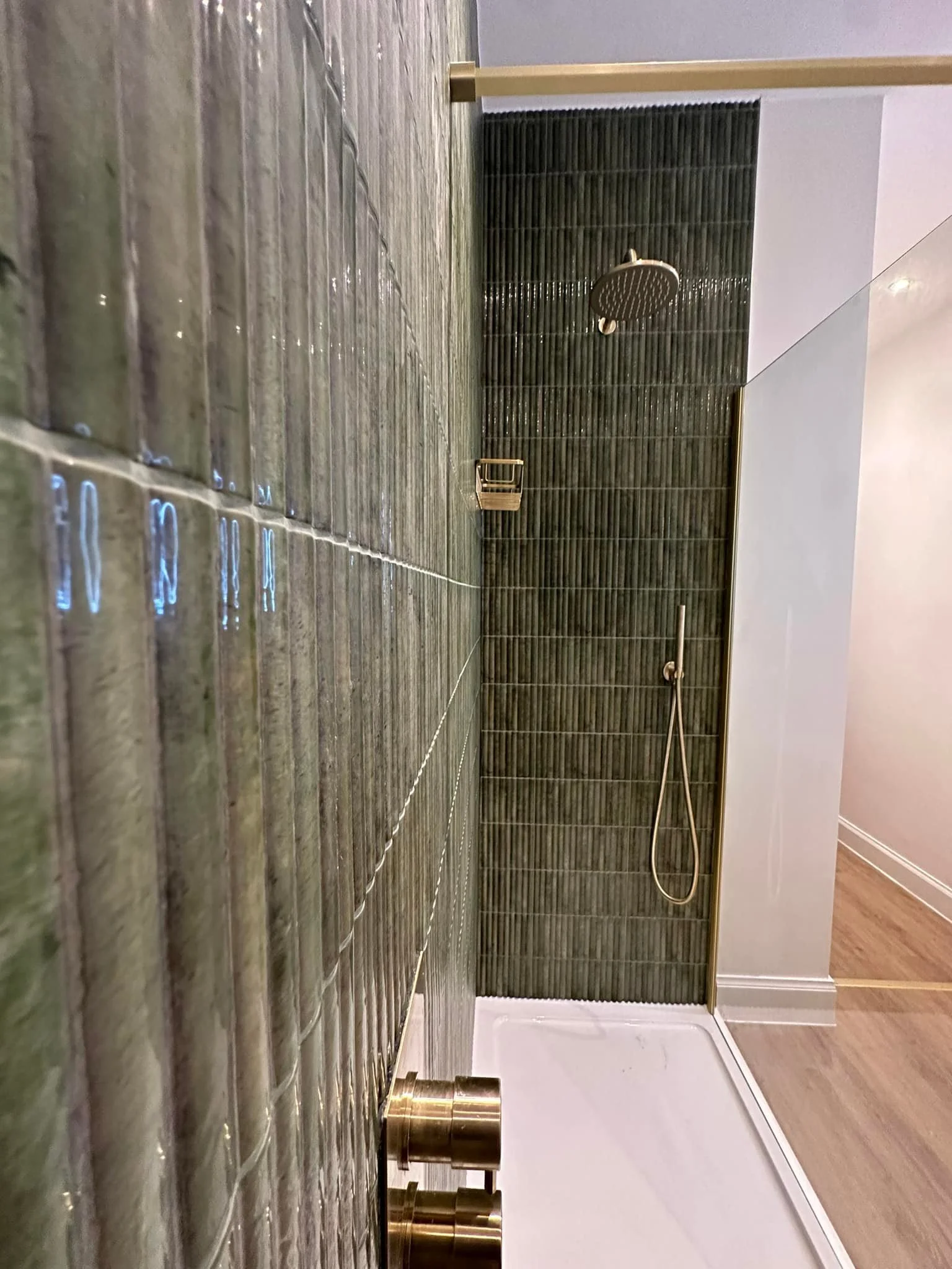 Modern bathroom shower with green textured tiles, a round showerhead, a handheld shower, and a small shelf, viewed from a low angle.