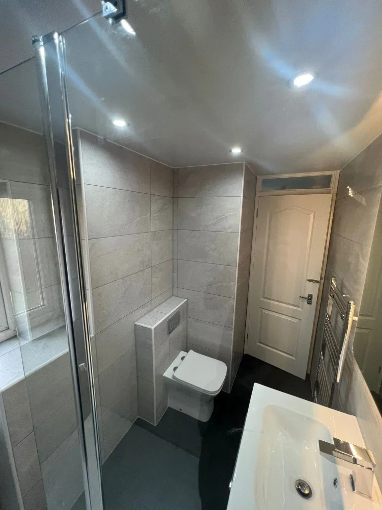 Compact modern bathroom with a shower stall, toilet, white sink, and heated towel rack with tiled walls and ceiling lights.