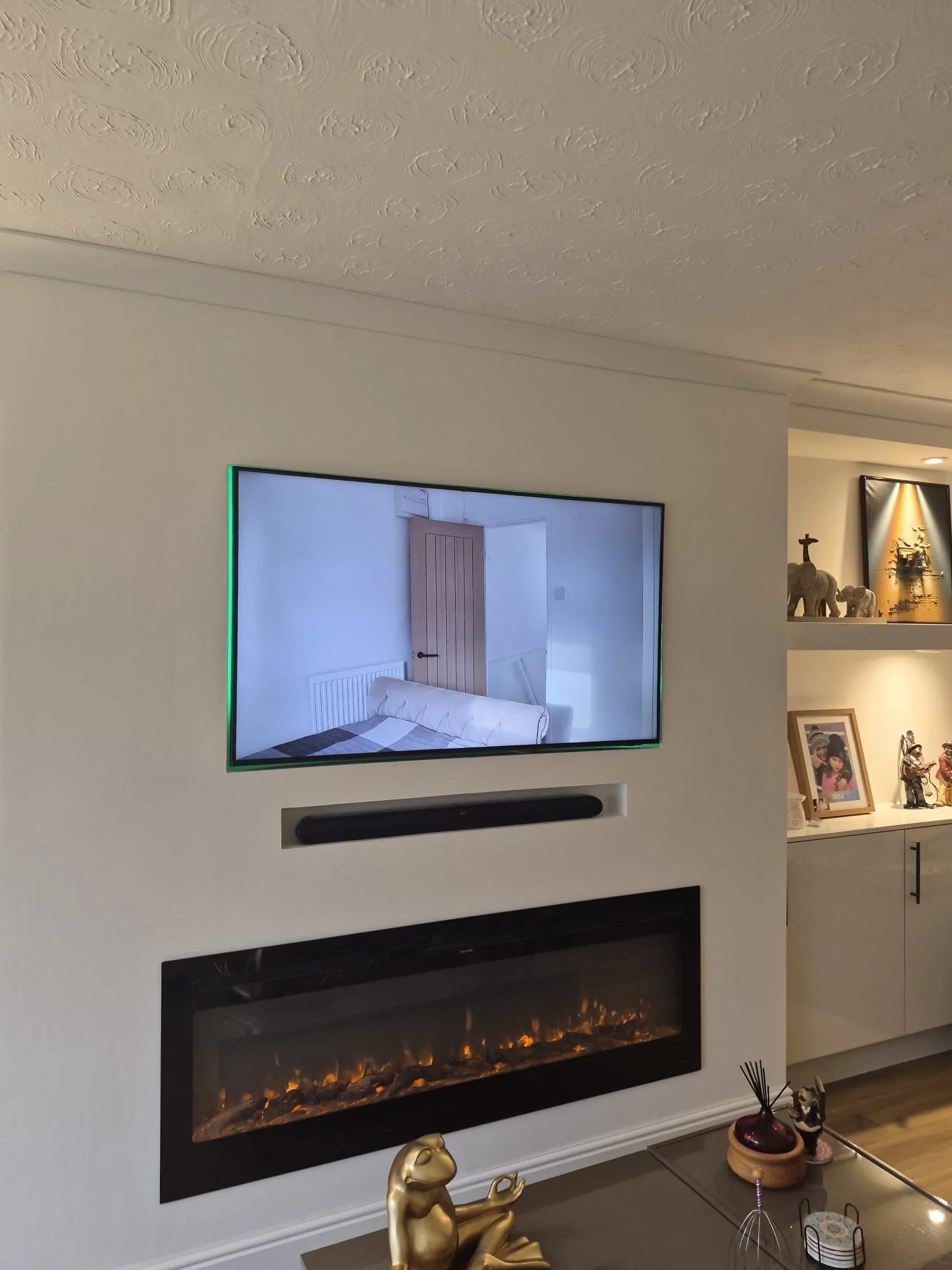 Living room with a mounted flat-screen TV above an electric fireplace with decor items on a table below, and a built-in shelf with decorative objects and framed photos on the right.