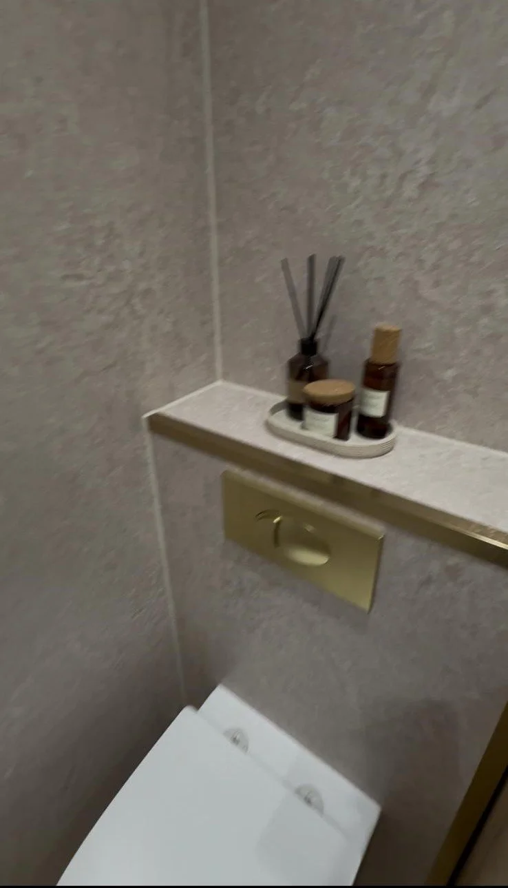 A bathroom corner with a small shelf holding diffuser sticks and various toiletry bottles, with a concealed toilet flusher behind a gold panel.