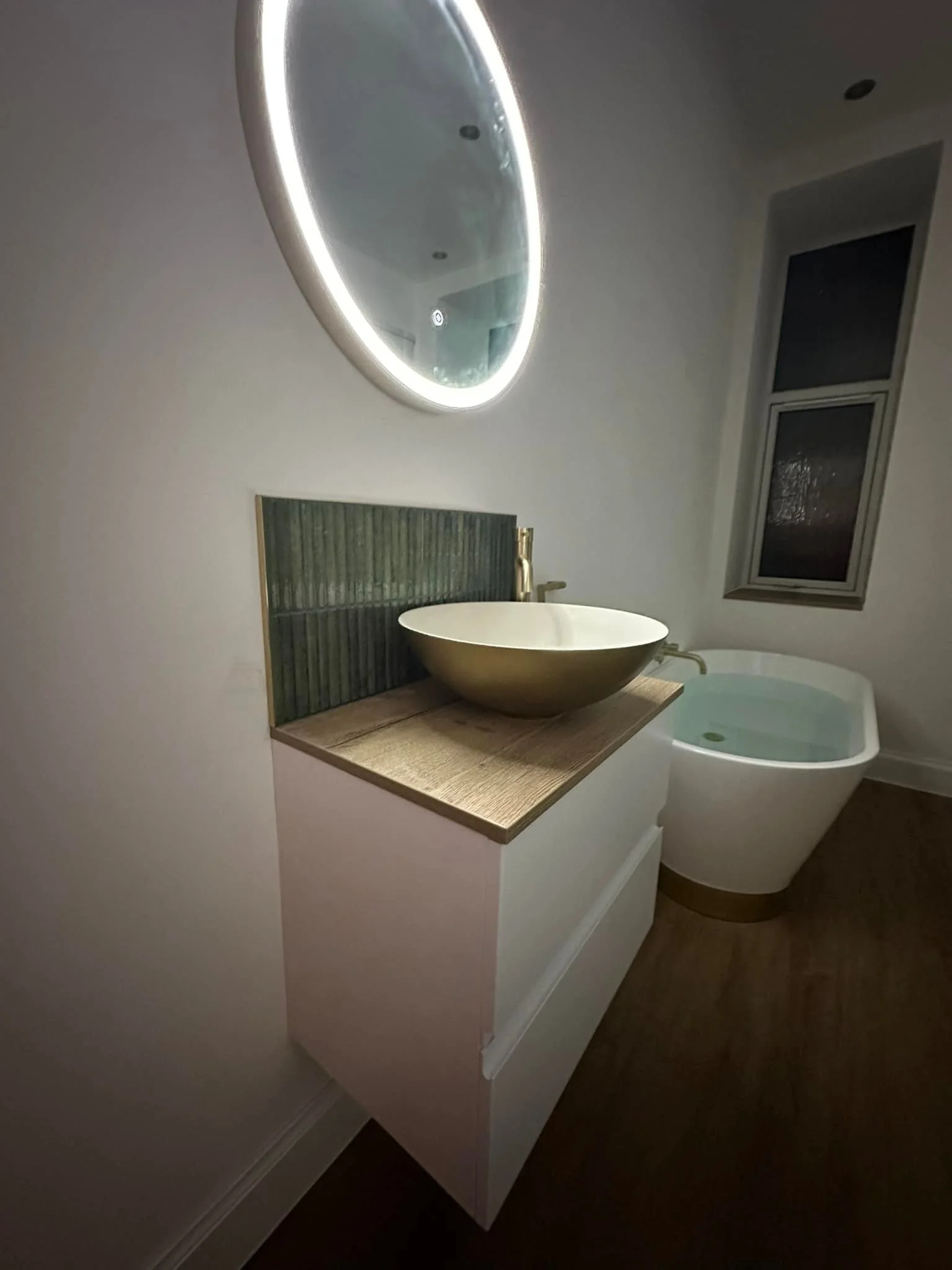 Modern bathroom with an oval backlit mirror, green backsplash, wooden vanity, golden vessel sink, and a free-standing bathtub with water in it near a window.