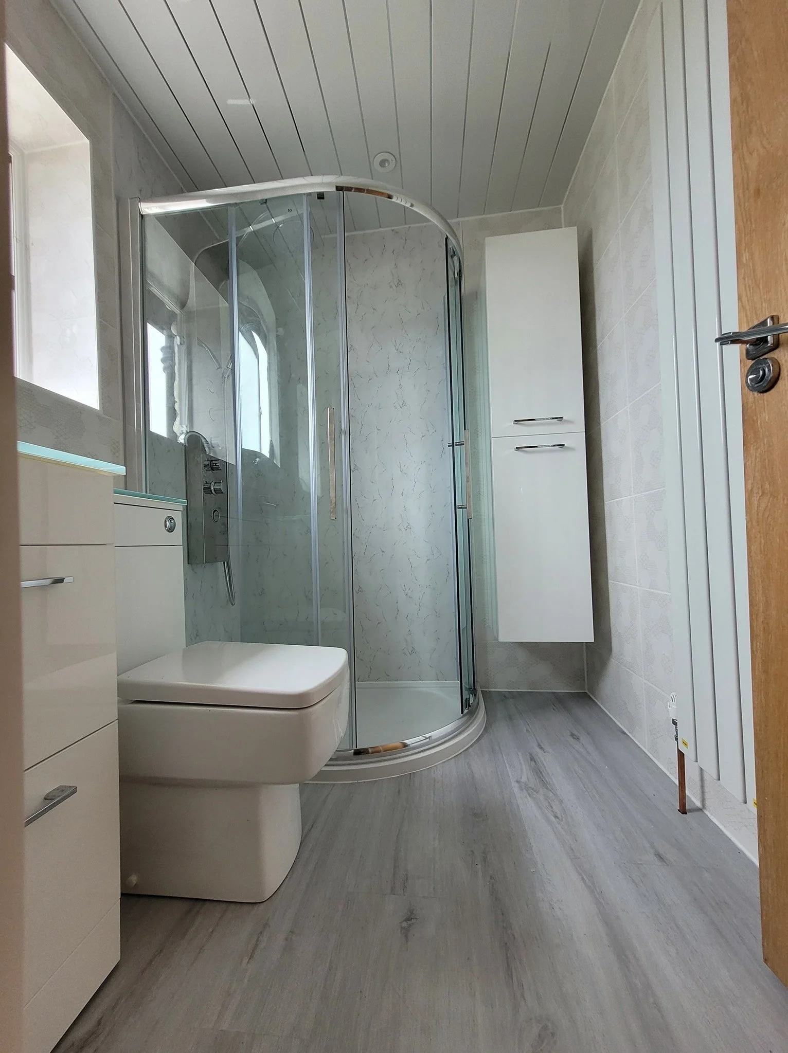 Beige bathroom with a standing shower stall, toilet, and white cabinets, with light wood flooring and a wooden door.