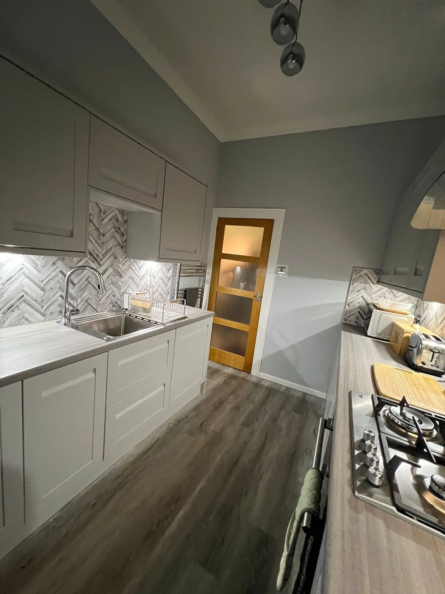 A modern kitchen with gray cabinets, a white countertop, a backsplash with a herringbone tile pattern, a stainless steel sink, and a gas stove. There are kitchen appliances and utensils on the counter and a wooden door with frosted glass panels leadi