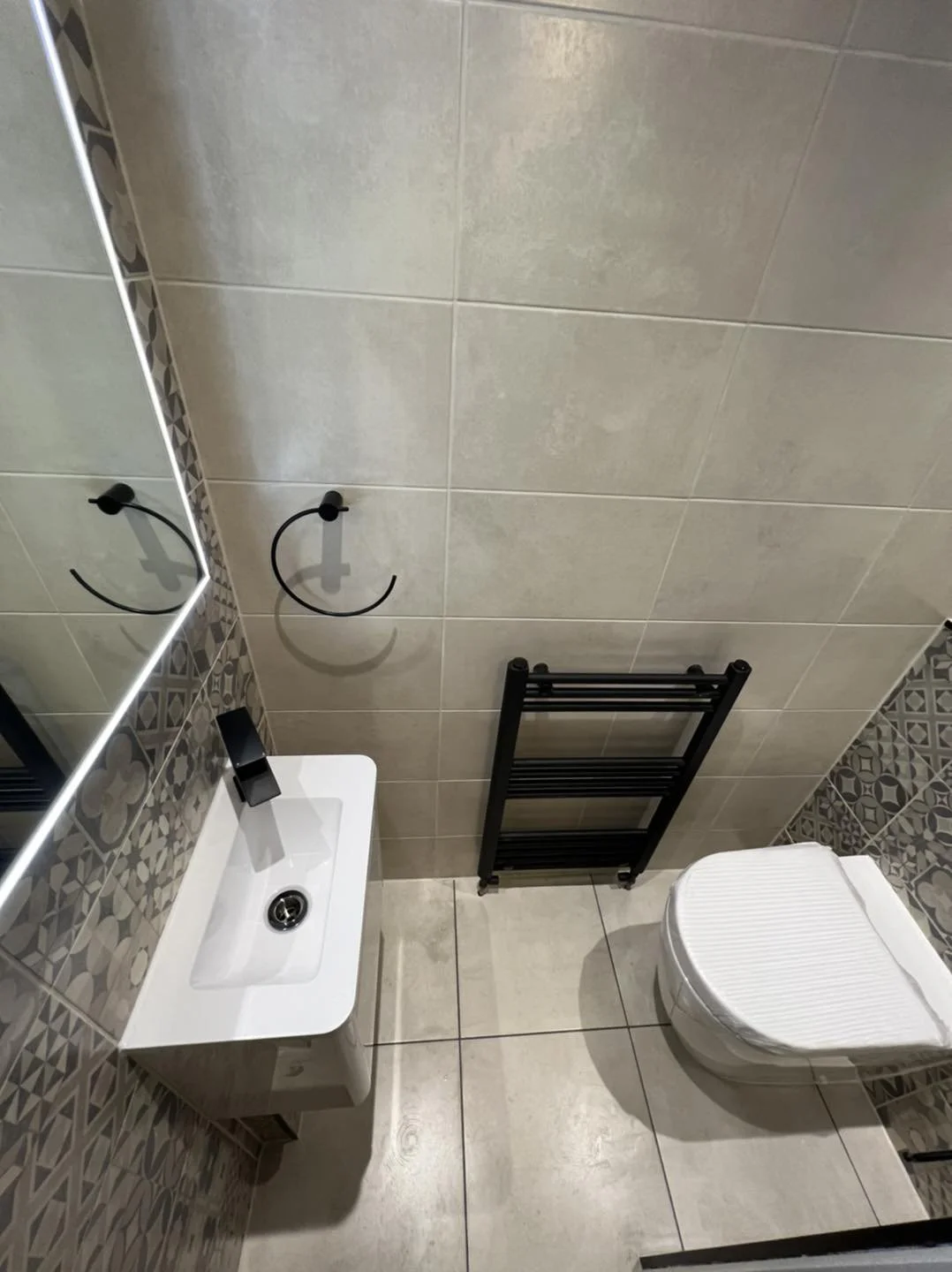 Modern bathroom with beige tiled walls and floor, a white sink with a black faucet, a mirror, two black towel rings, a black heated towel rail, and a white toilet with a lid.