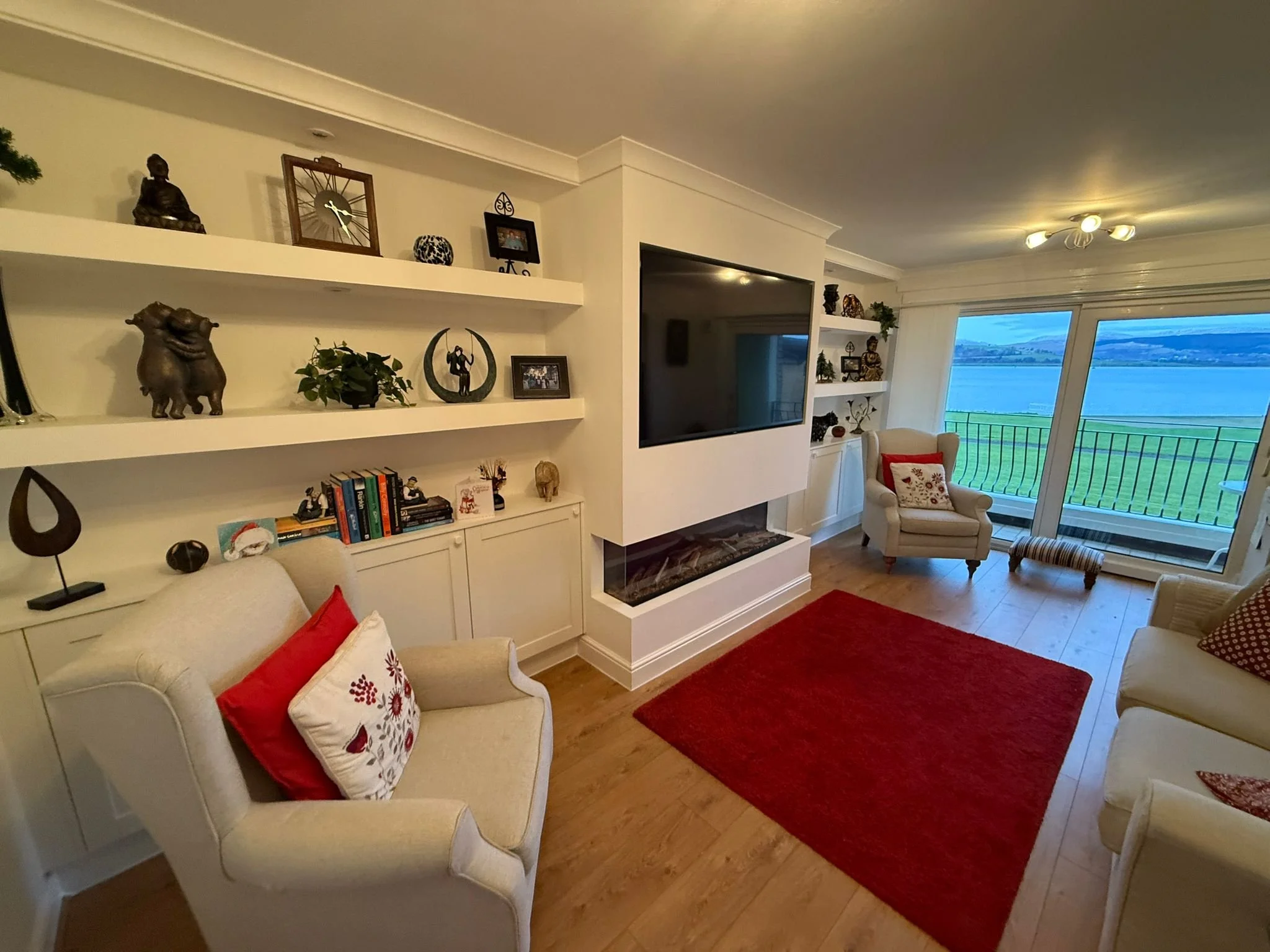 Living room with white furniture, red accents, built-in bookshelves with decorative items, flat-screen TV, large windows with a view of a green field and water, and a fireplace.