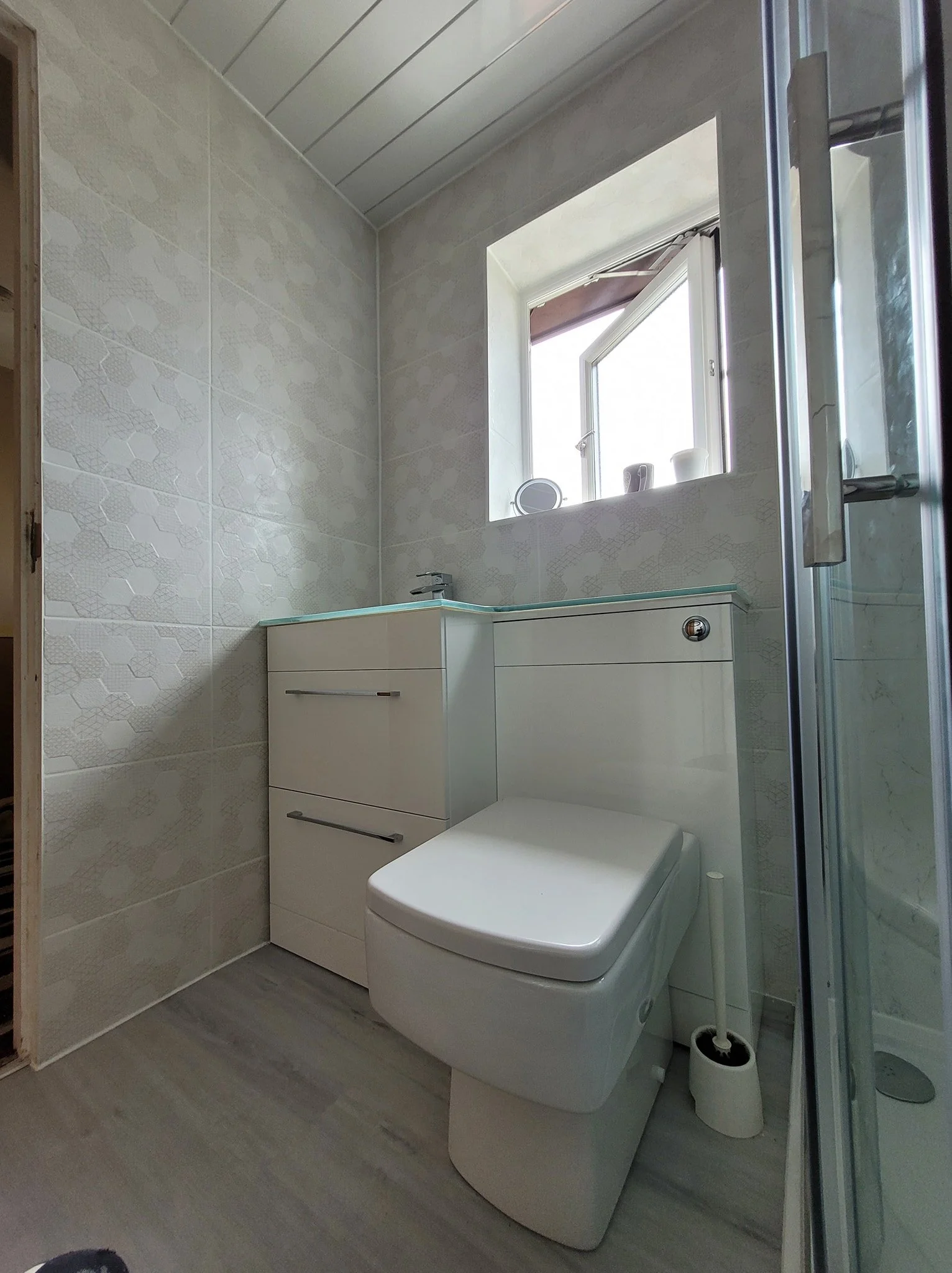 A small bathroom featuring a modern toilet, a white cabinet with drawers, a small sink, and a window with a shelf holding three cups. The walls are tiled with a subtle floral pattern, and a glass shower enclosure is partially visible on the right.