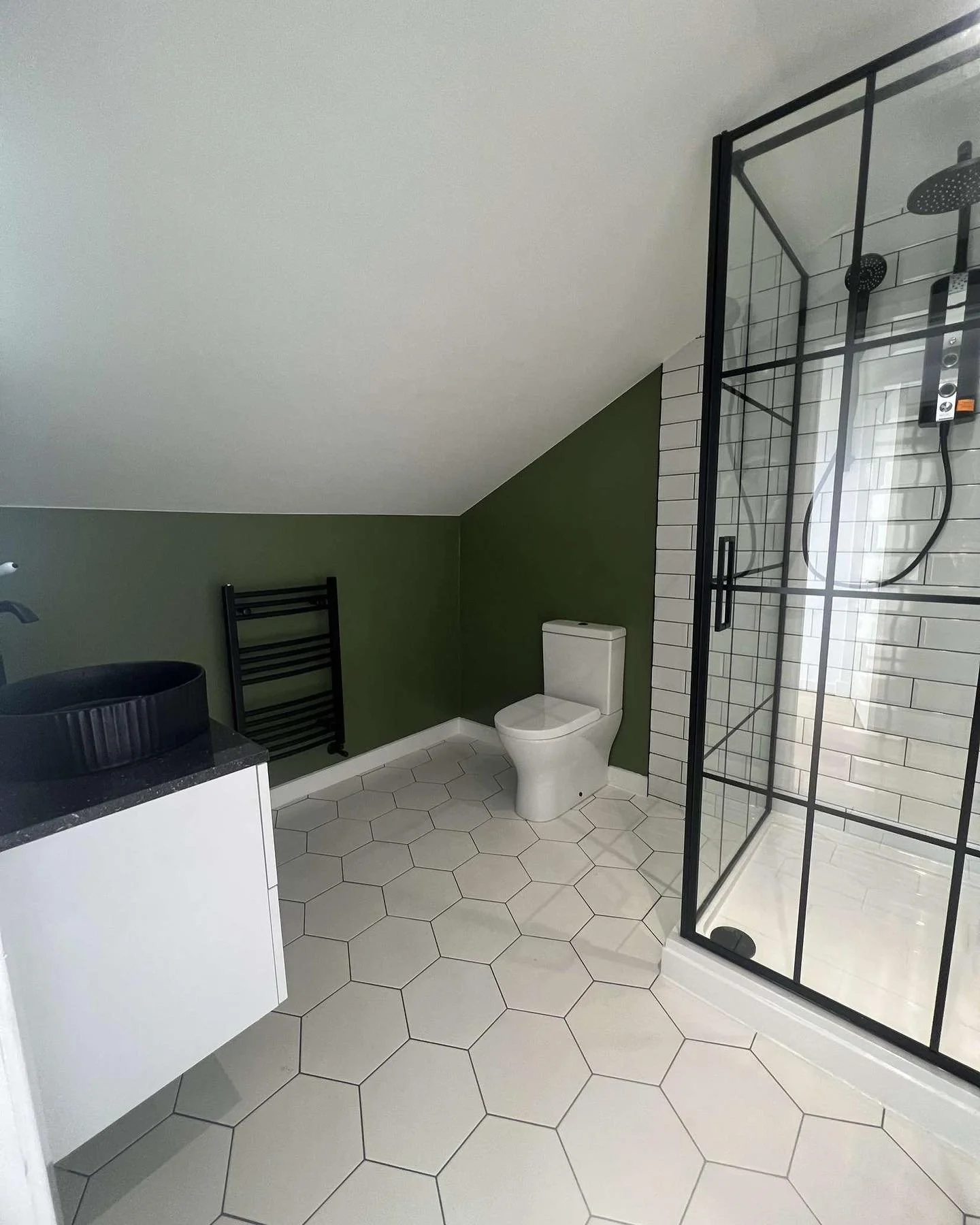 Modern bathroom with white hexagonal floor tiles, green accent wall, white toilet, black towel warmer, black vessel sink on a white vanity with black countertop, and glass shower enclosure with black framing and white subway tiles inside.