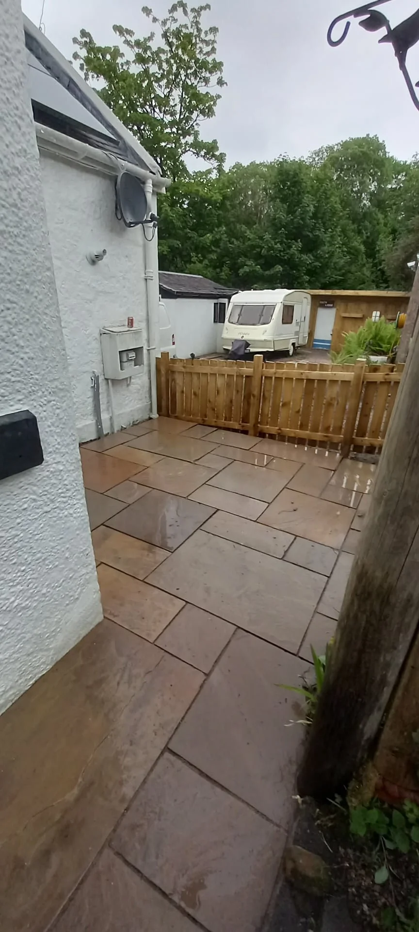 Wet outdoor patio with beige stone tiles, small wooden fence, caravan trailer parked behind the fence, neighboring house with an air vent, green trees in the background, rainy sky.