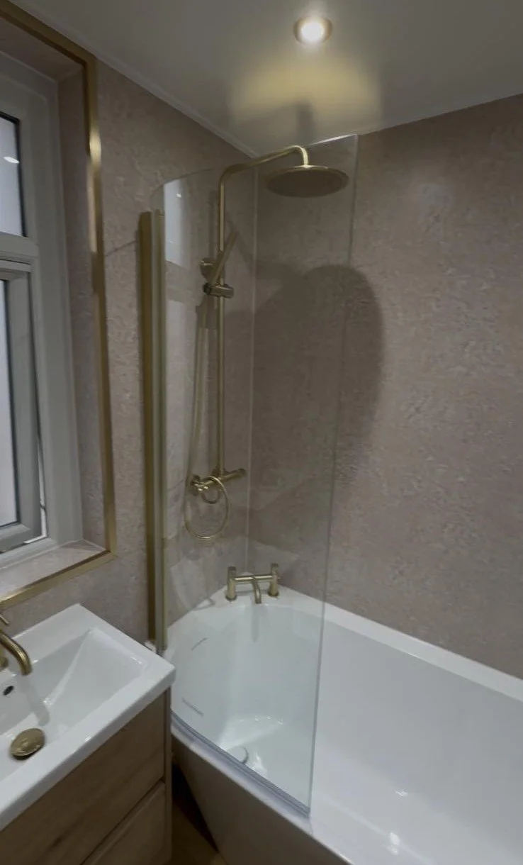 A bathroom with a bathtub, a glass shower partition, a showerhead, a small white sink with a gold faucet, a mirror, and a window. The wall has beige textured tiles, and the ceiling has a recessed light.