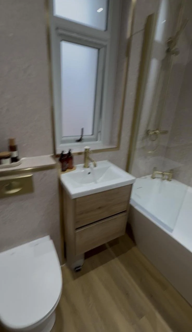 Small bathroom with a white toilet, wooden vanity with a sink and gold faucet, window above the sink, and a bathtub with a gold faucet and showerhead. The walls are tiled beige and the floor is wood-patterned.