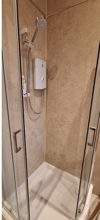 A small, enclosed shower with beige tiled walls, a white electric shower unit, and a glass door with metal handles.