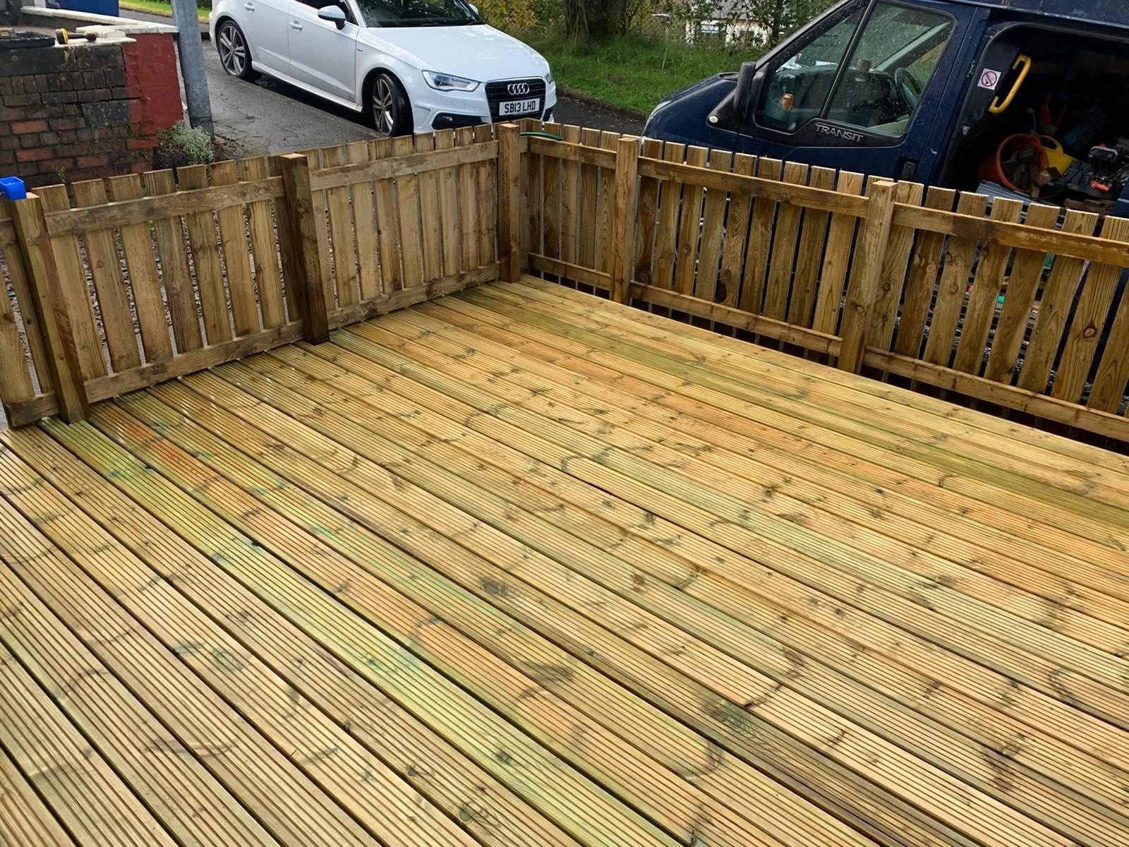 An outdoor wooden deck with a new wooden fence and clean wooden floorboards, with parked cars and a garden in the background.
