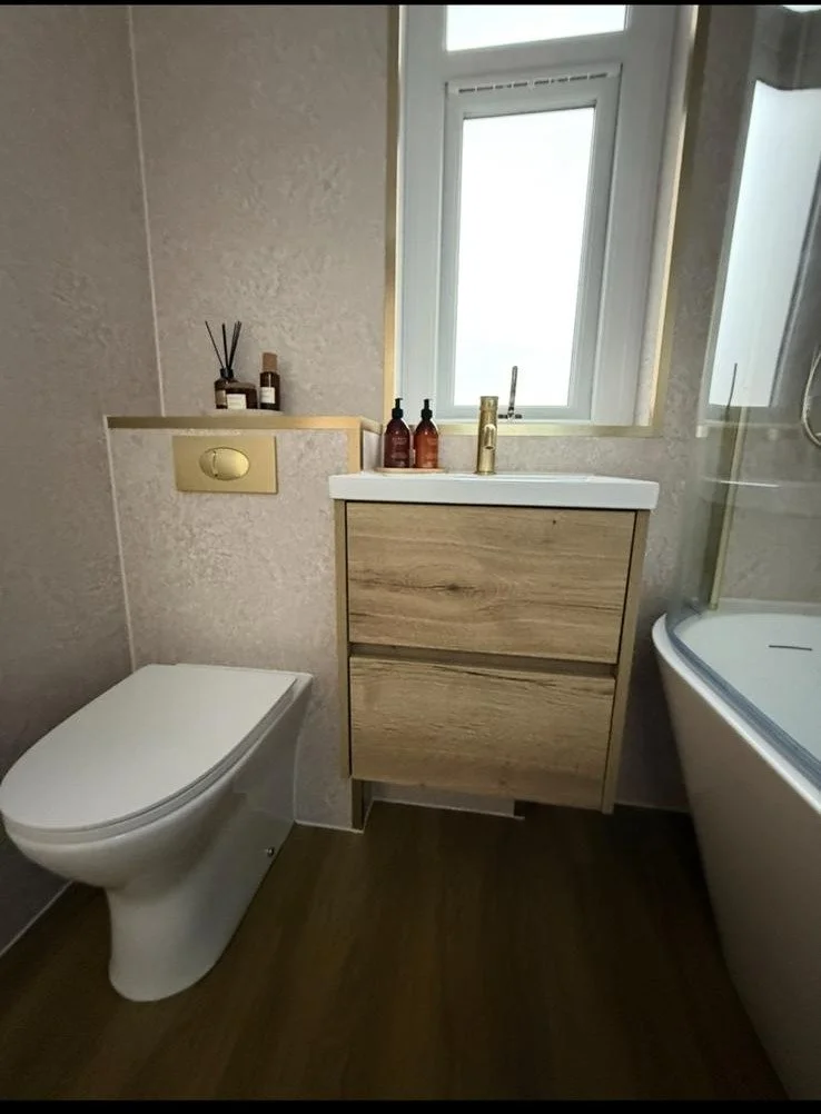 A modern bathroom with a wall-mounted toilet, a wooden vanity with a sink, and a curved bathtub with a glass shower enclosure. There are bottles on the vanity and a small window above.