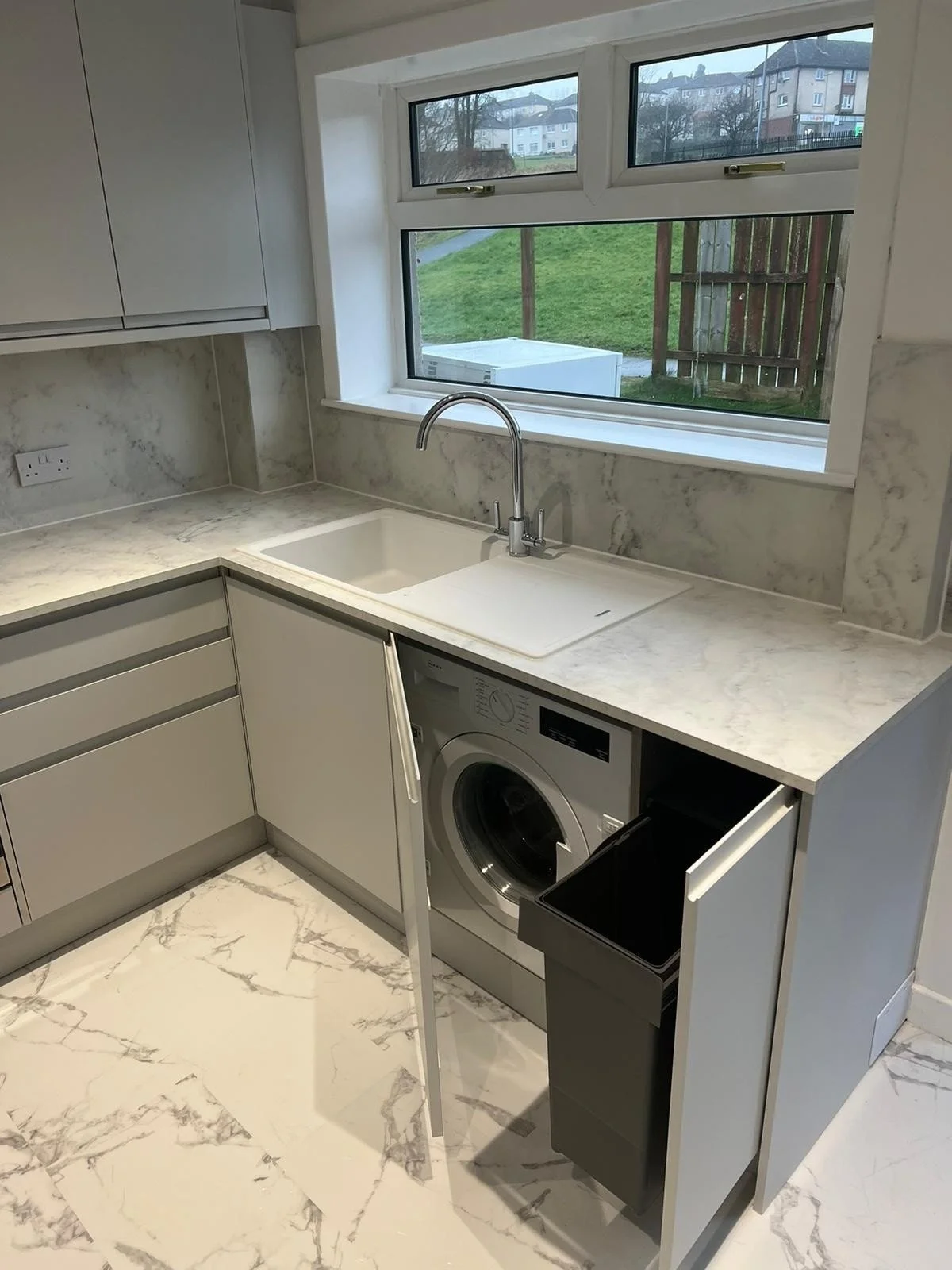 Modern kitchen with white marble countertops, a stainless steel sink, a washing machine, and large windows overlooking a green yard and neighboring houses.
