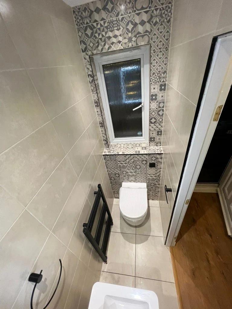 Small bathroom with beige tiled walls and floor, a window with patterned tiles around it, a wall-mounted toilet, and a black towel rack leaning against the wall.