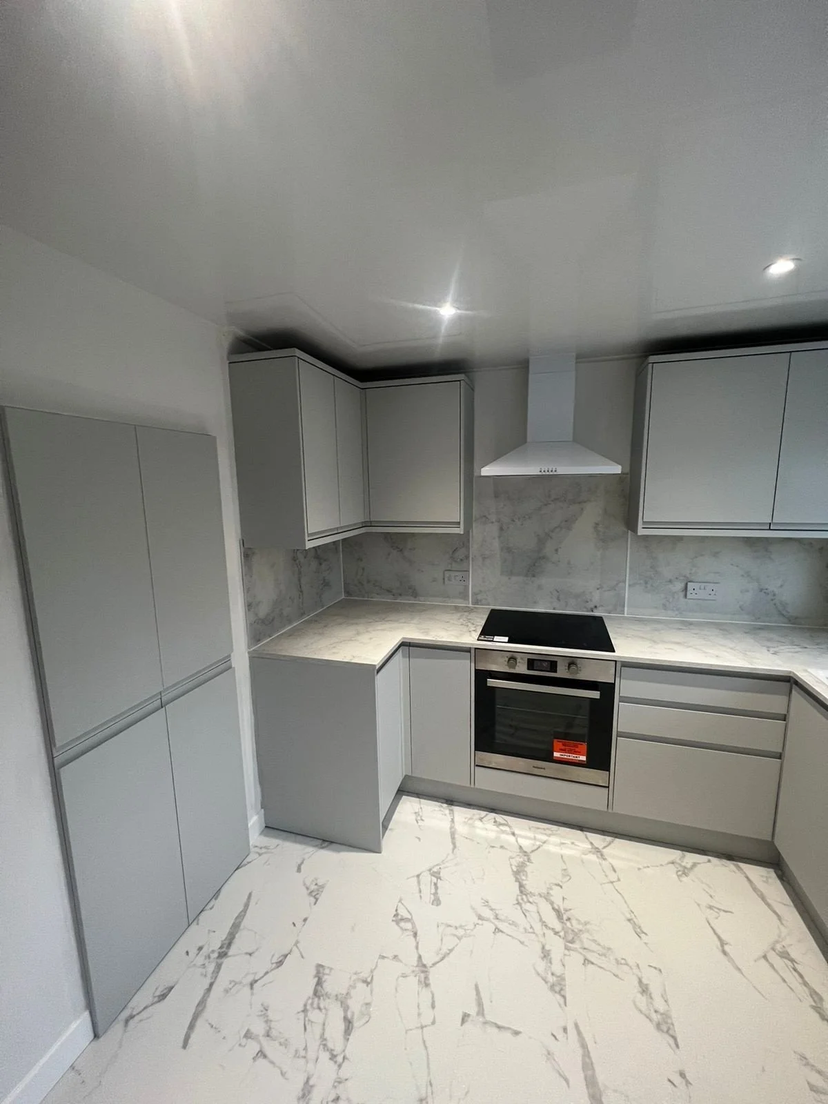 Modern kitchen with light gray cabinets, marble countertops, marble backsplash, and a marble floor. There is an oven, a range hood, and electrical outlets.