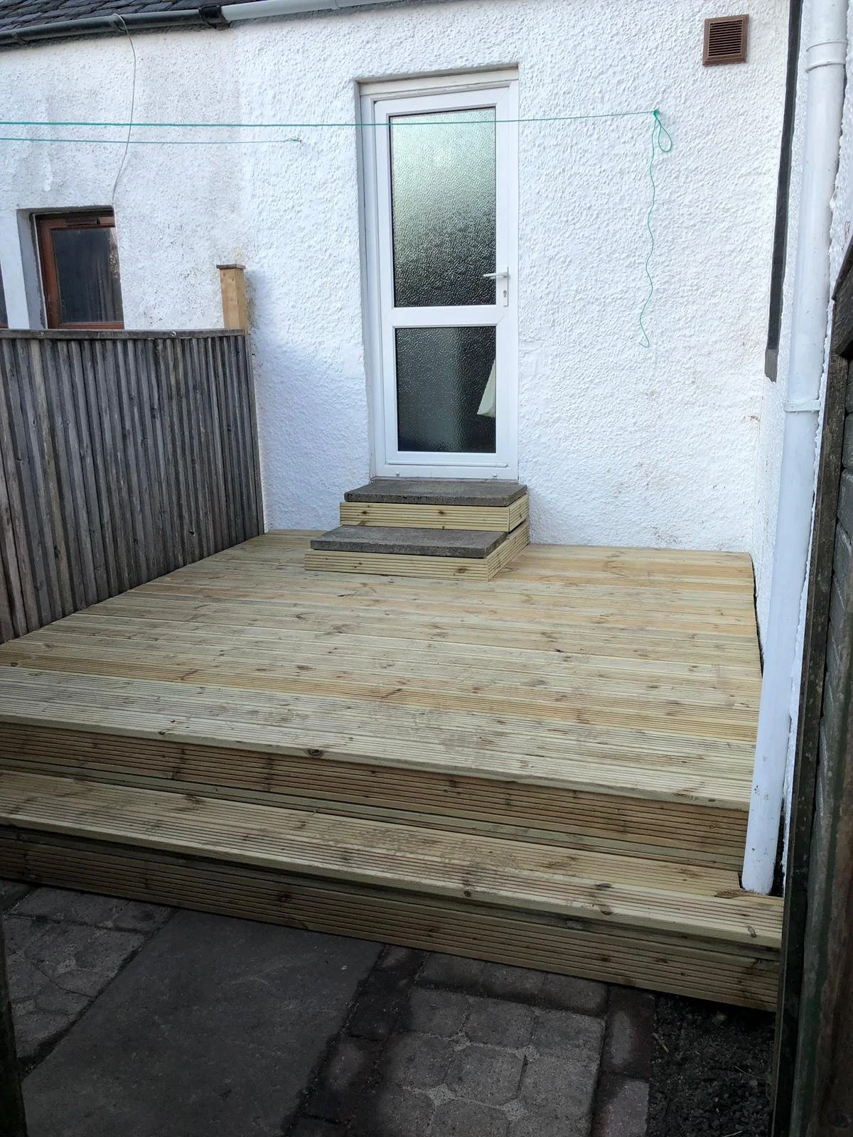 Freshly built wooden deck with stairs next to a white back door, bordered by a wooden fence on the left and a brick wall on the right.