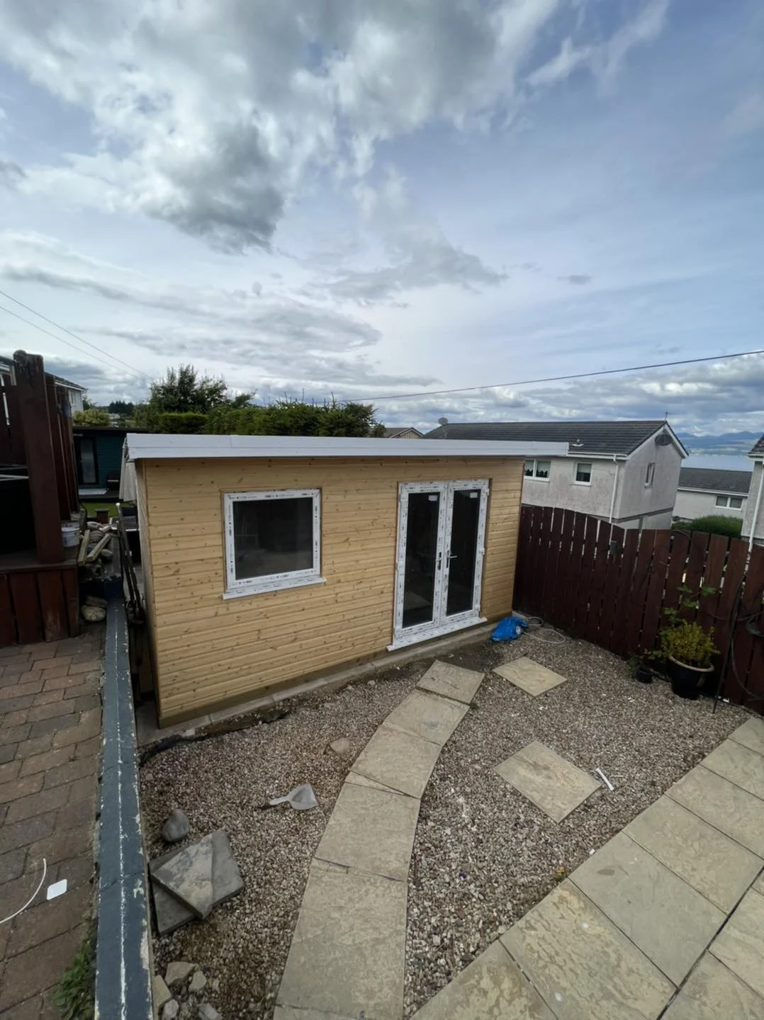 View of a small wooden shed with French doors and a window in a backyard, with a gravel and stone path, surrounded by a red wooden fence and neighboring houses under a cloudy sky.