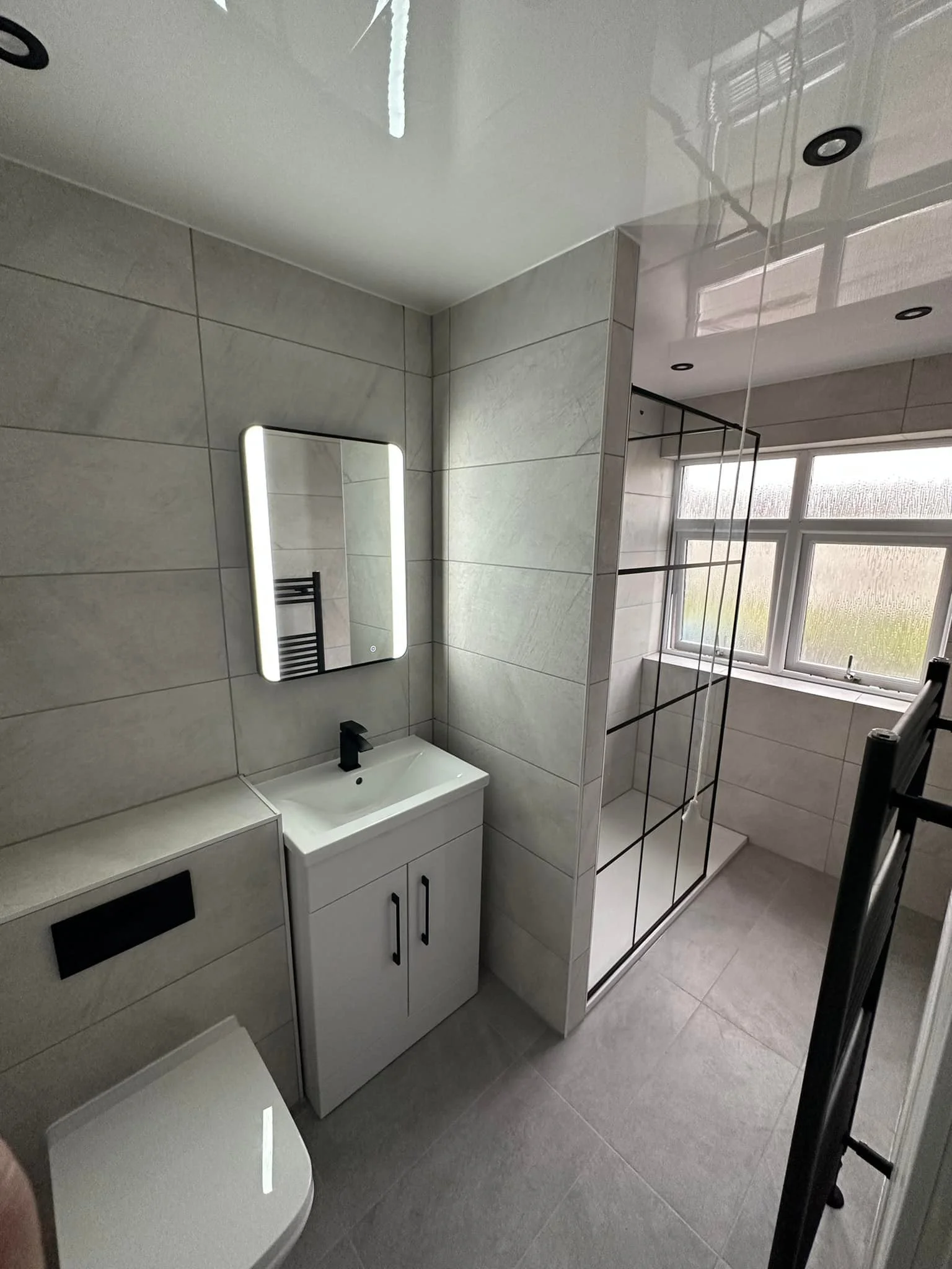 Modern bathroom with a small vanity, illuminated mirror, walk-in shower with glass door, window, and grey tiled walls and floor.