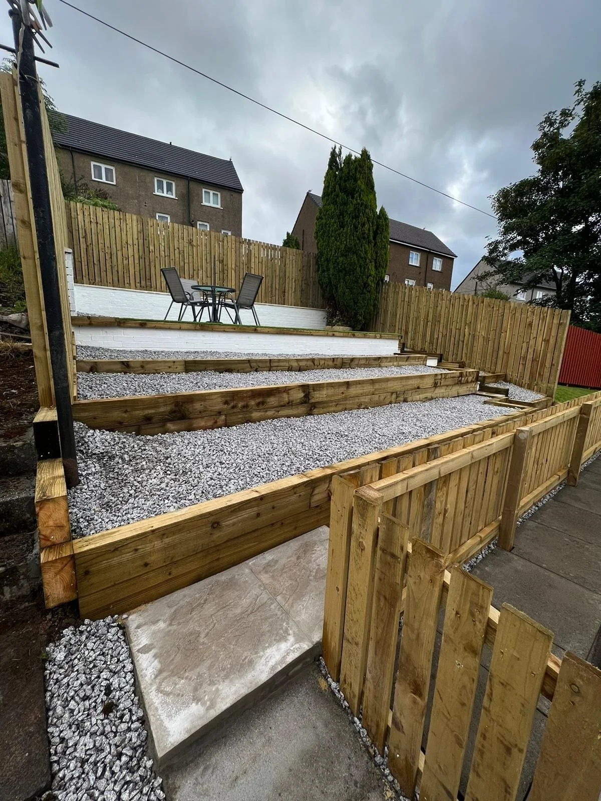 A tiered backyard landscape with wooden retaining walls, gravel ground cover, a small concrete pathway, a patio table with four chairs, and a tall evergreen tree surrounded by fencing, under a cloudy sky.