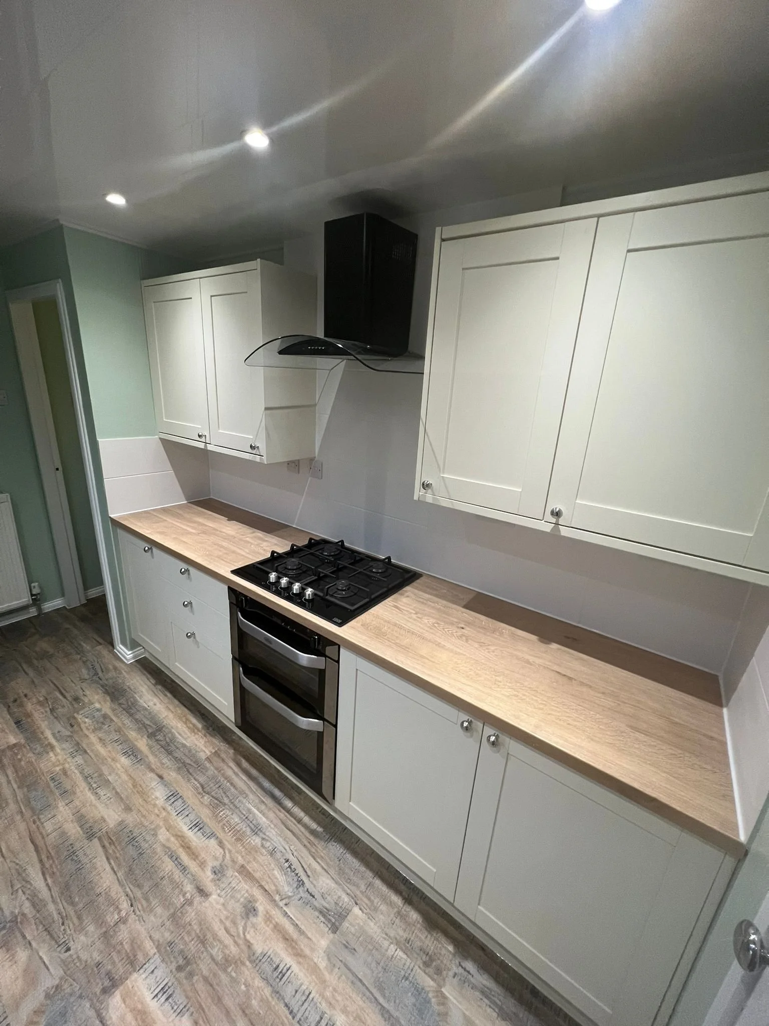 Modern kitchen with white cabinets, wooden countertop, gas stove, oven, black range hood, and laminate flooring.