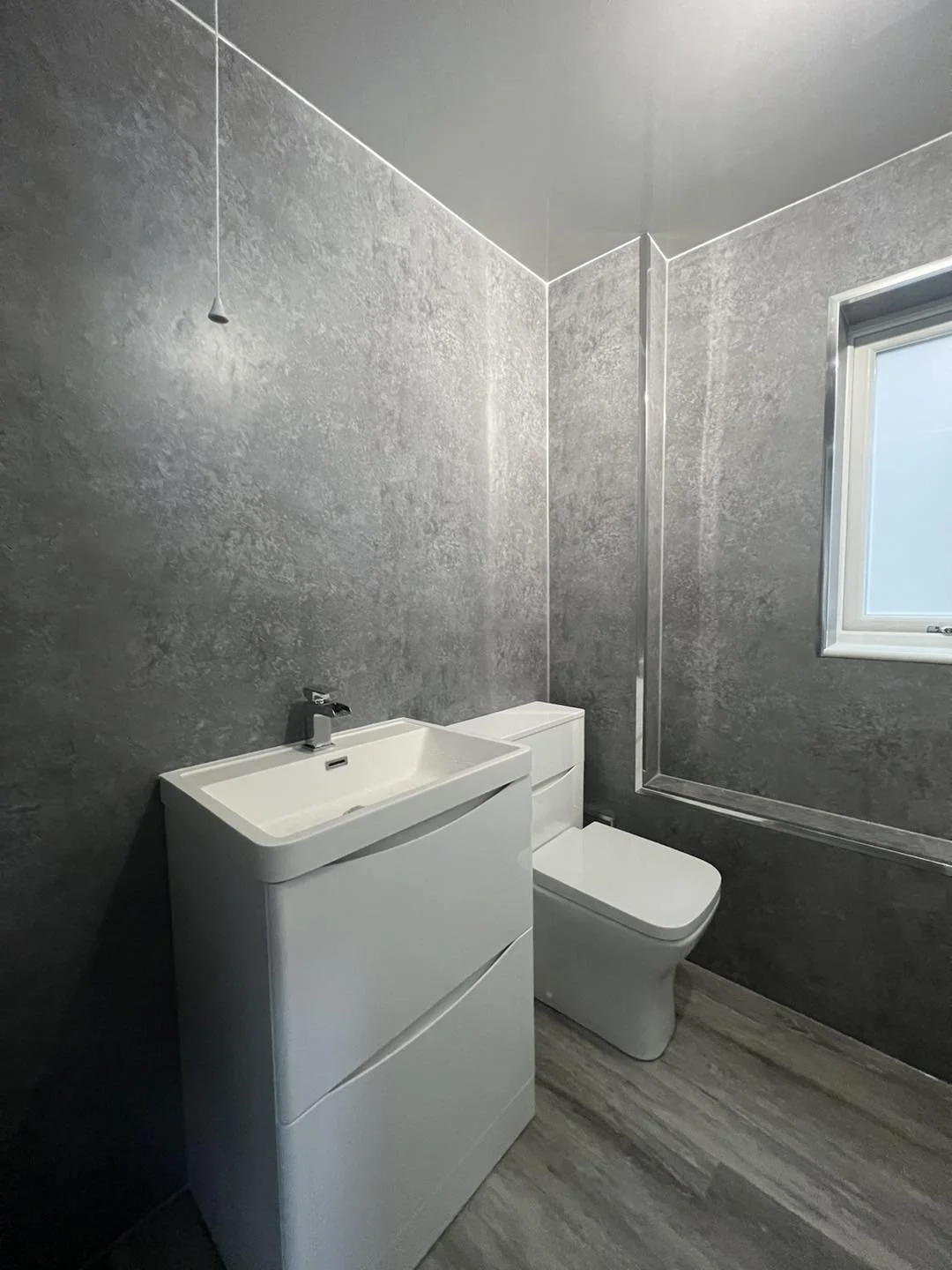 A modern bathroom with gray textured walls, a white sink with a cabinet, a white toilet, and a window on the right side.
