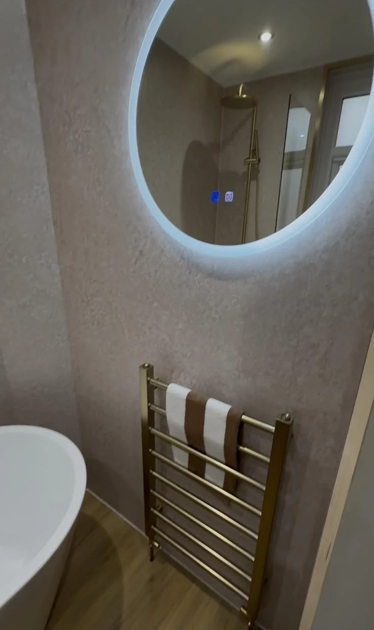 Bathroom with a round backlit mirror, a modern gold towel rack with a white and brown towel, a bathtub, and a walk-in shower with a gold showerhead, in a room with beige walls and wooden flooring.