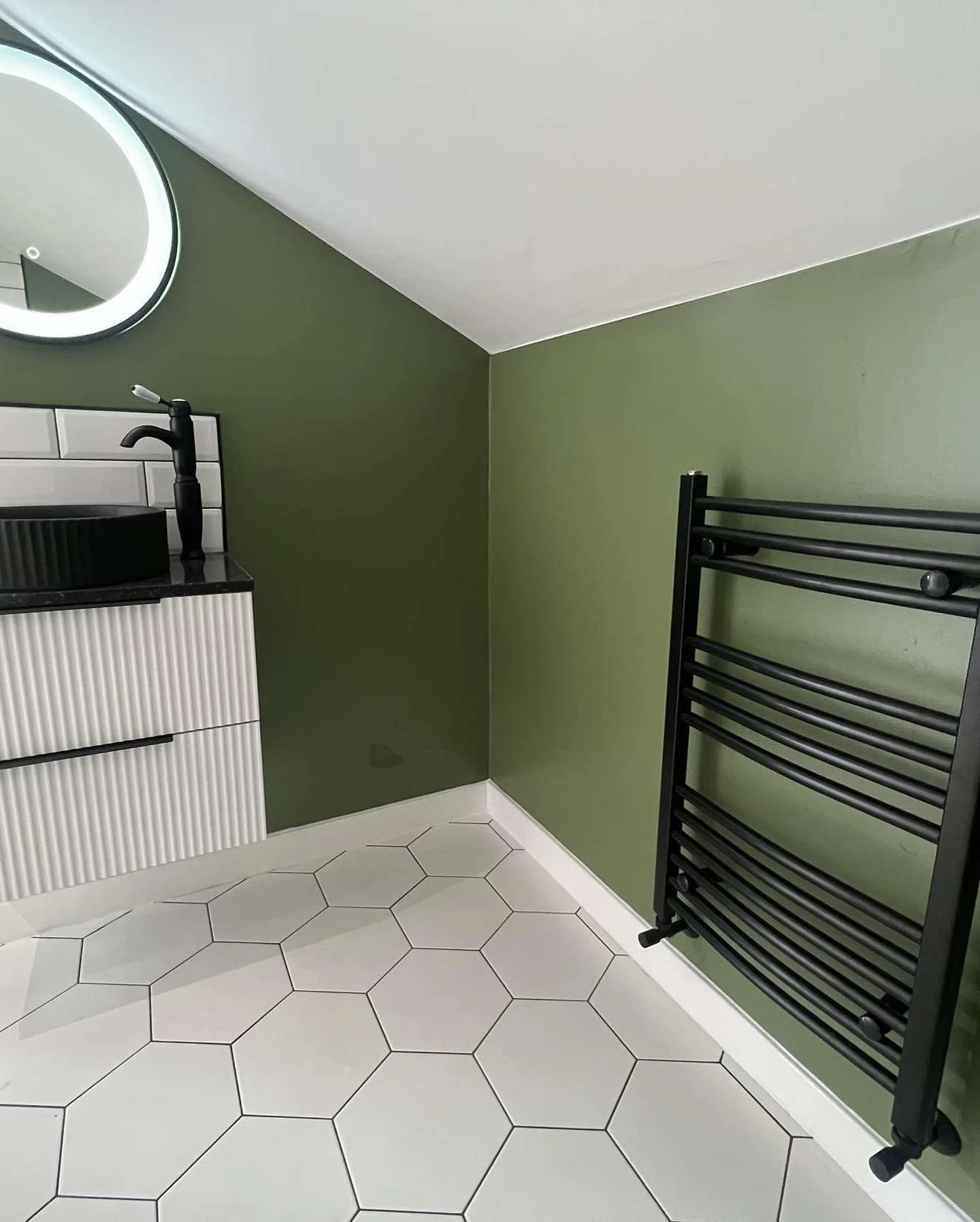 A bathroom corner with a black towel radiator, a small vanity with a black sink and faucet, green walls, white hexagonal floor tiles, and a round mirror with a light.
