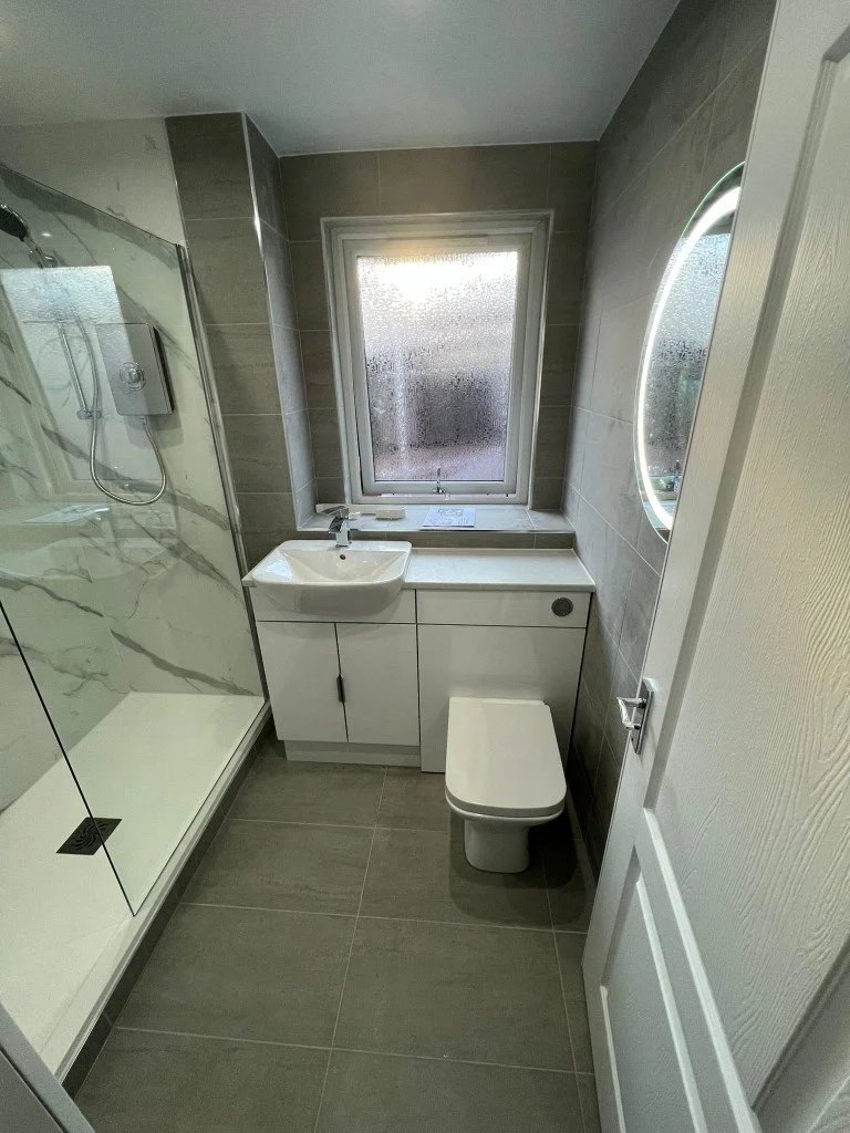 Small modern bathroom with a walk-in shower, a white vanity with a sink, a frosted window, a circular mirror, and a toilet, with gray tiles on the floor and walls.
