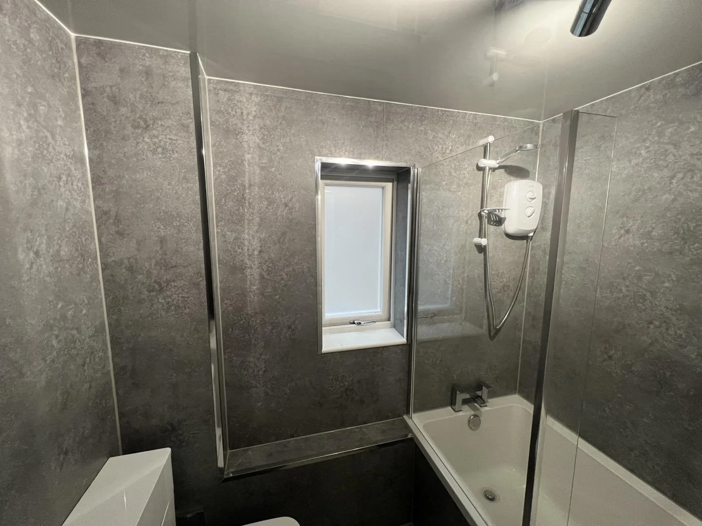 Small bathroom with gray marble walls, a bathtub with a handheld showerhead, a window above the bathtub, and a white ceiling light.