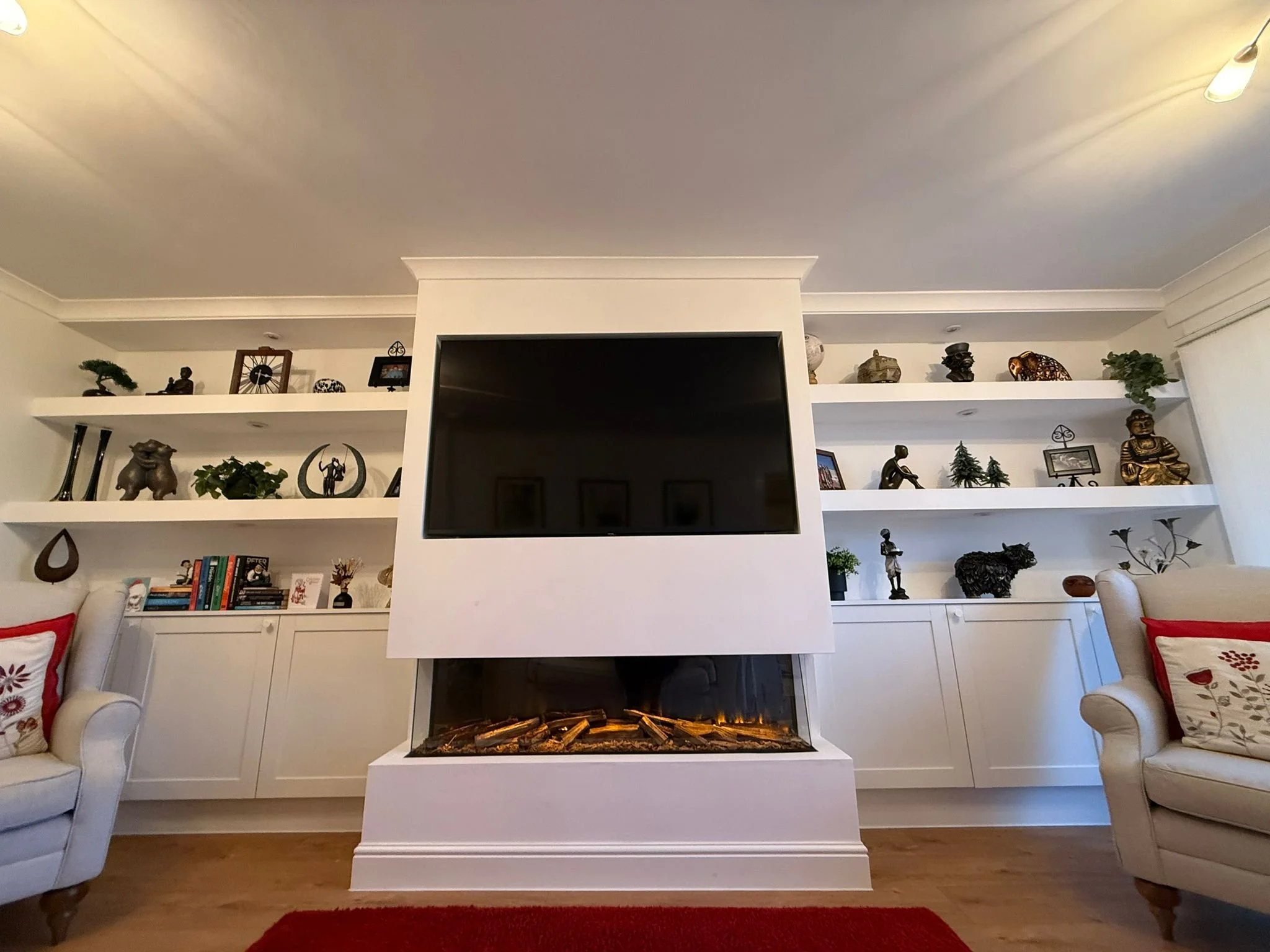 Living room with white built-in bookshelves, a mounted flat-screen TV, and a modern fireplace below it. There are two beige armchairs with red and white throw pillows, and a red rug on hardwood floors. Decor items on the shelves include books, plants