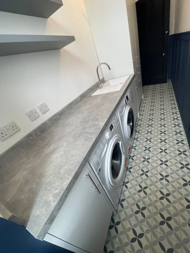 Laundry room with a gray countertop, a small sink, a faucet, and two front-loading washing machines. The floor has black, gray, and white patterned tiles.