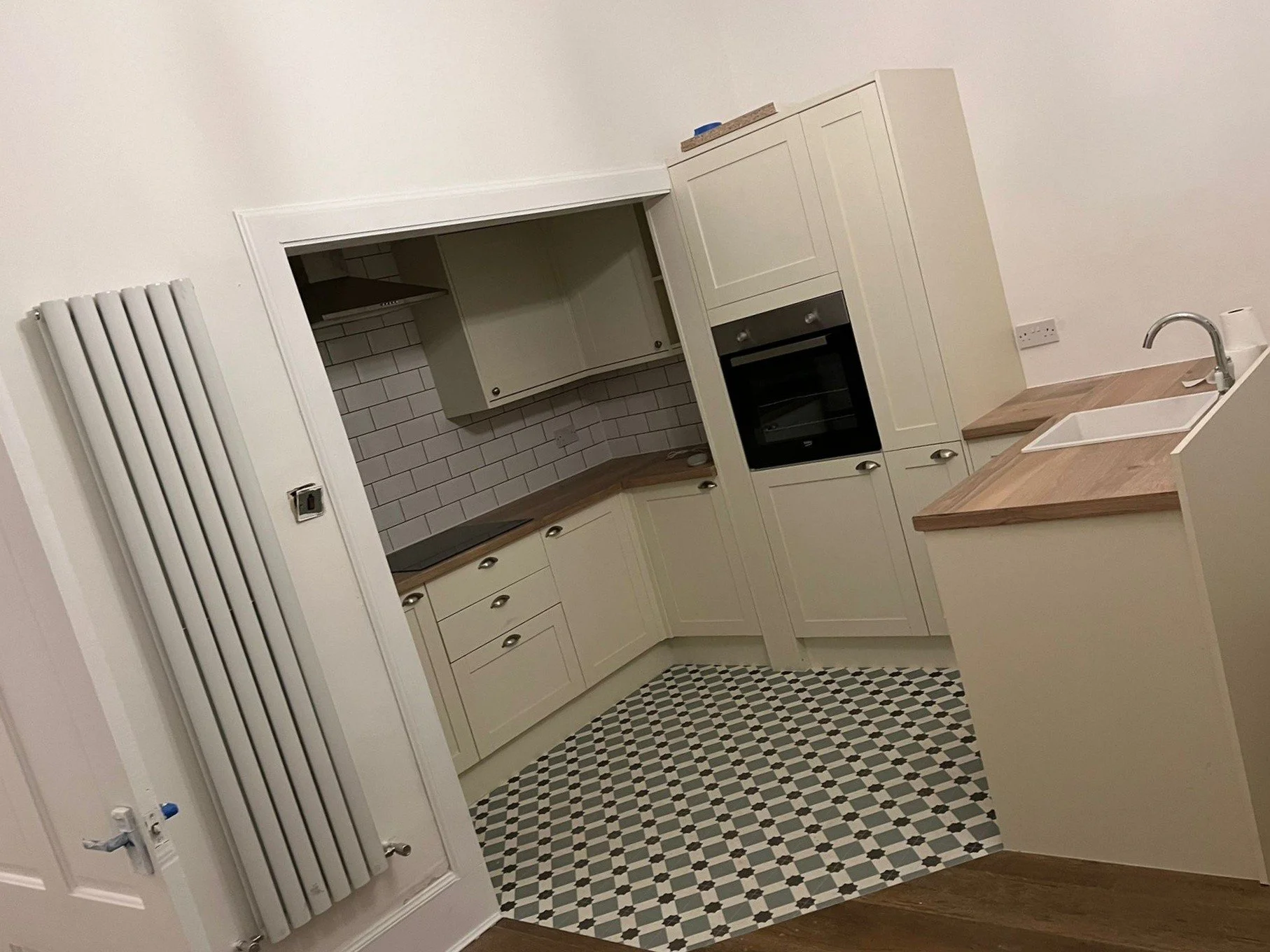 Interior view of a kitchen with white cabinets, wooden countertops, a built-in oven, white subway tile backsplash, a white sink, and a geometric patterned black and white floor.