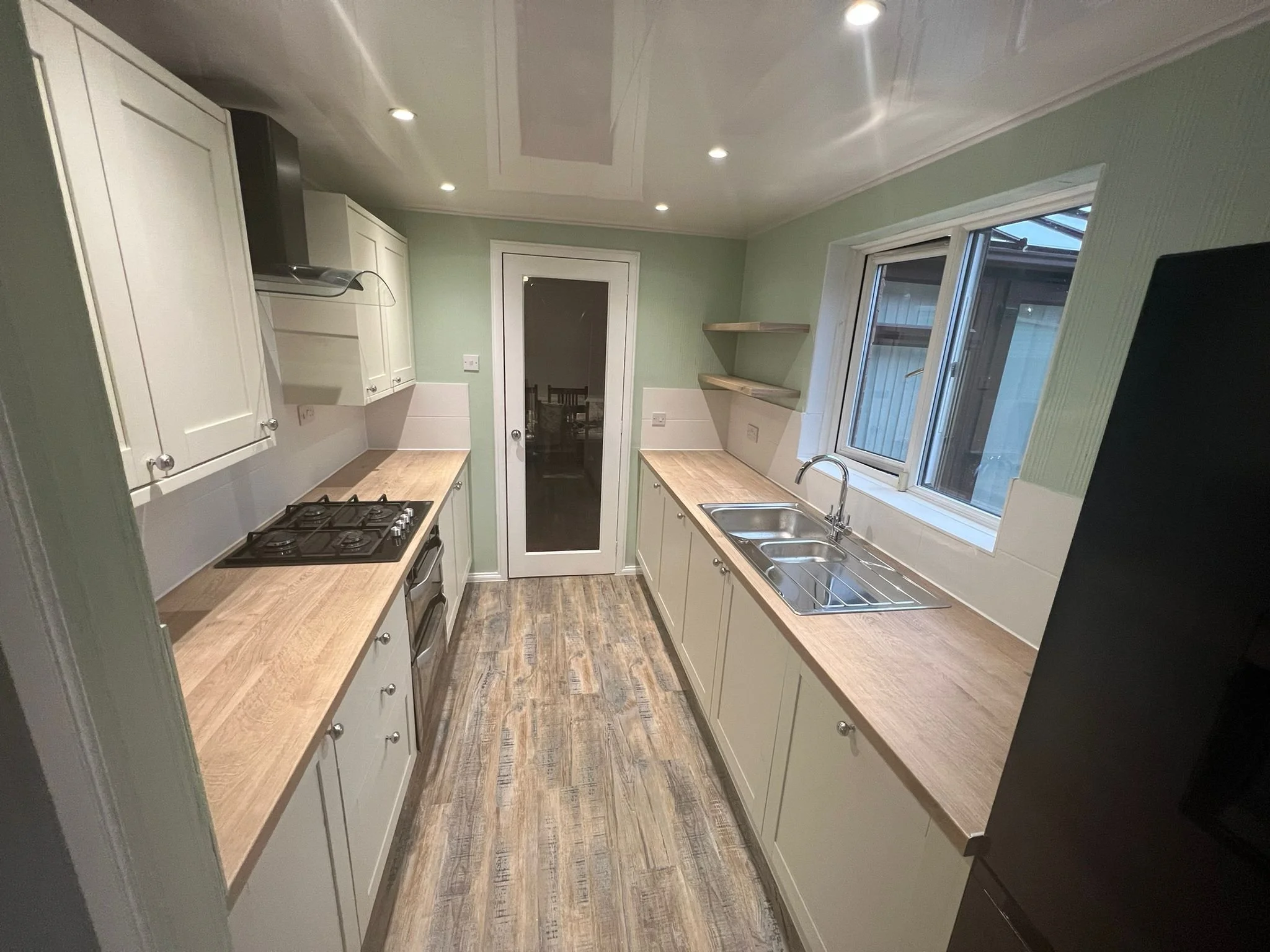 A modern kitchen with cream-colored cabinets, wooden countertops, a stainless steel sink, a gas stove, and a black refrigerator, with a window and shelves on the right wall.
